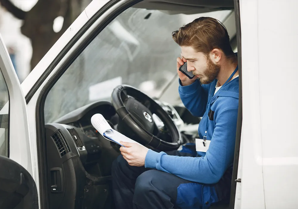 A worker on a mobile sat in a van with the door open, looking at documents on a clipboard